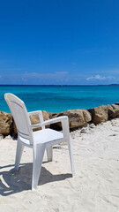 A white plastic chair placed on a pristine white sand beach, facing the expanse of a crystal-clear turquoise ocean. Can be used to represent relaxation, tropical vacations, solitude, an idyllic escape