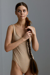 young woman with long braided hair wearing beige bodysuit thoughtfully looking aside against plain...