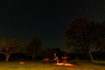 Fototapeta premium Woman sitting by backyard fire pit looking up at the stars in the night sky above