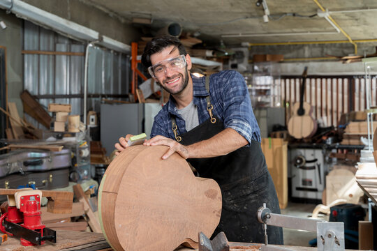 Smiling male carpenter sanding wooden guitar body in workshop, wearing apron and safety goggles for protection