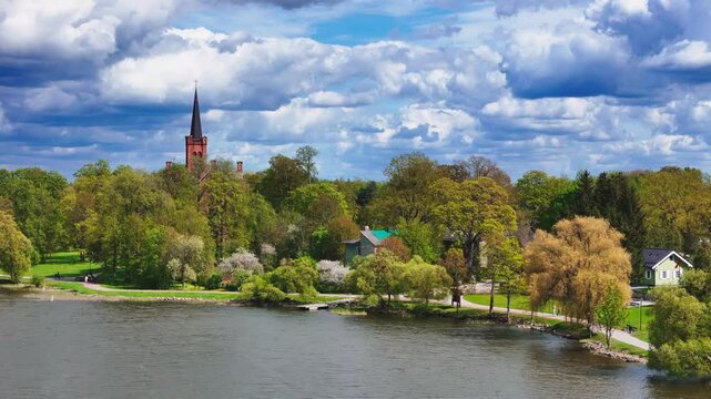 idyllic spring day in Vaxjo, Sweden, captured from an elevated wide-angle view, showcasing vibrant green trees, a distant red-brick church spire, and fluffy white clouds reflected in the calm lak