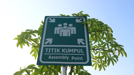 Emergency assembly point sign with pictogram and bilingual text "TITIK KUMPUL / Assembly Point", photographed under clear sky with green tree background