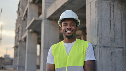 Smiling Engineer at Construction Site: A confident engineer, clad in a safety helmet and vest, radiates positivity while leaning against the structured architecture of a construction site.