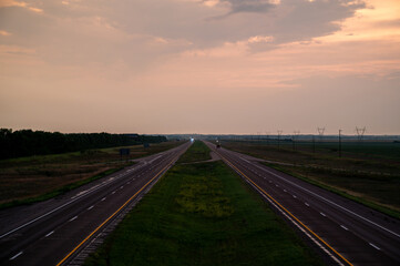 Fototapeta premium Wide view of empty highway stretching into the distance at dusk with soft clouds and gentle hues in the sky, surrounded by open fields and power lines