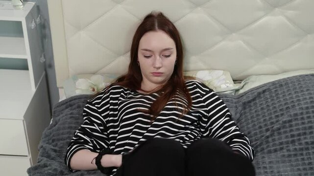 Teenager casually sitting on soft gray blanket, carefully manipulating small orange object with focused attention while resting comfortably on white bed