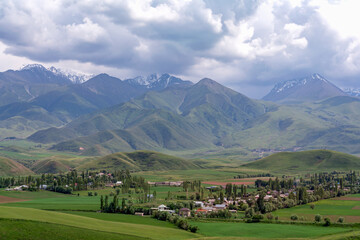 View from a high hill to the village, located in a green valley, and to the mountain peaks of different shades, some of which are covered in snow among the clouds.