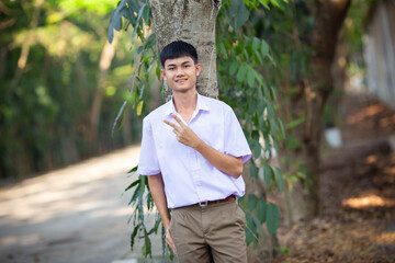 Thai boy student in official Thai school uniform (khaki or brown shorts), properly dressed, stand firmly in front of the tree with two fingers up, horizontal image
