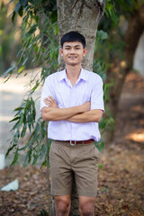 Thai boy student in official Thai school uniform (khaki or brown shorts), properly dressed, stand firmly in front of the tree with 
crossing his arms over his chest