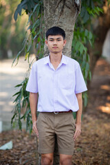 Thai boy student in official Thai school uniform (khaki or brown shorts), properly dressed, stand firmly in front of the tree with his thumbs in pockets