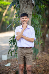 Thai boy student in official Thai school uniform (khaki or brown shorts), properly dressed, stand firmly in front of the tree with arms crossed, and one hand under his chin