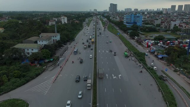 Aerial perspective of a wide highway with light traffic, lined by green patches and scattered houses under a cloudy sky.