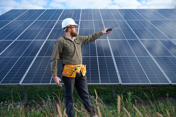 Engineer in uniform with protective helmet near solar station holding walkie talkie. Supervisor is...