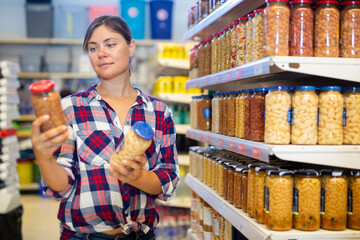 Interested young woman standing near shelves with jars of canned vegetables in supermarket, choosing beans in sauce