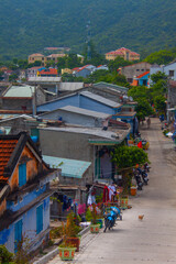 Fototapeta premium Colorful Vietnamese village street with traditional houses, scooters, laundry hanging, and mountains in the background. A small dog walks along the quiet road. 