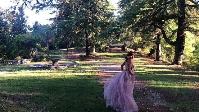Romantic slow motion of young European woman in powder pink open-back maxi dress running through magical green forest. Dreamy cinematic aesthetic and fairytale atmosphere.