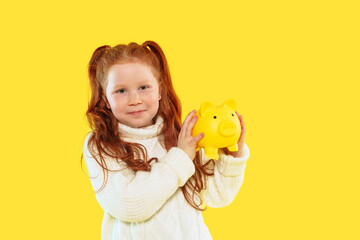 Child holds yellow piggy bank against bright yellow background with cheerful expression while promoting the idea of saving money