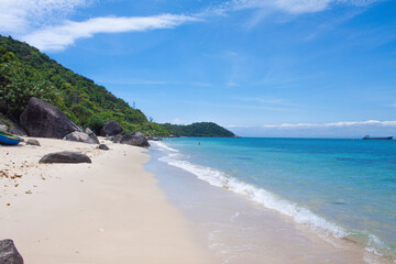 Tropical beach scene with turquoise waves gently washing ashore, a lone swimmer in the sea, and distant islands under a clear blue sky. 