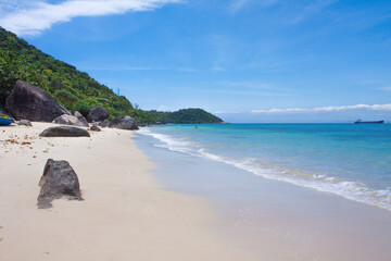 Tropical beach scene with turquoise waves gently washing ashore, a lone swimmer in the sea, and distant islands under a clear blue sky. 
