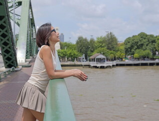 Asian woman with short, dark hair stands by a green railing on a bridge, gazing thoughtfully at the...