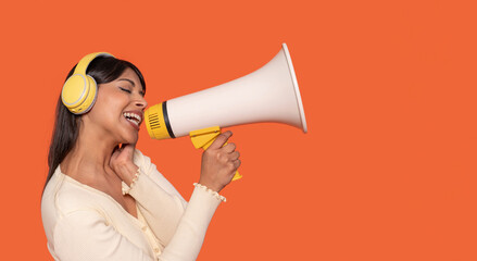 Energetic young woman wearing headphones passionately sings into a large megaphone against an orange background during a lively performance