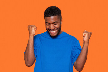 Joyful young man celebrates success with raised fists against a vibrant orange background, expressing happiness and excitement in a casual blue shirt