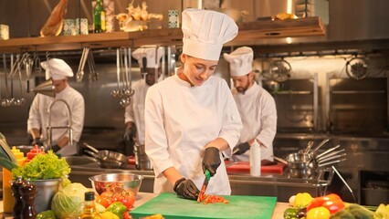 Portrait of cheerful successful skillful female chef chopping tomato on cutting board with knife....