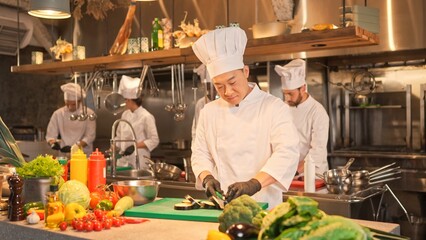 Focused skillful professional Asian chef cooking delicious salad chopping aubergine on cutting board with knife. Concentrated male cook wearing white uniform and black gloves in kitchen.
