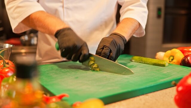 Close-up view of professional skillful male chef's hands chopping cucumber on cutting board. Hard-working experienced caucasian cook preparing ingredients before cooking delicious food.