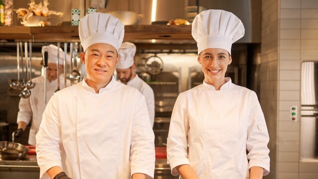 Two attractive professional cooks cutting and chopping vegetables with knife at modern kitchen of restaurant. Beautiful caucasian female and skillful asian male chefs talking and cooking.
