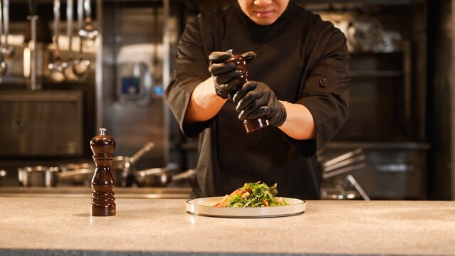 Portrait of cheerful professional asian male chef in black uniform and gloves adding salt and pepper to delicious salad. Attractive skillful happy cook smiling enjoying cooking at kitchen.
