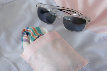 Stylish featuring a pastel-colored men’s shirt with a folded pocket square in soft tones. Transparent-frame sunglasses with dark lenses rest on the fabric, creating a refined and summery fashion.
