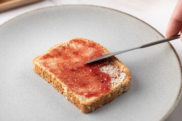 Close-up of whole grain toast with strawberry jam on a gray plate, male hand in action and neutral background. Concept of simple food and natural eating