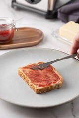 Close-up of a man's hand spreading strawberry jam on whole grain bread over white marble, with breakfast utensils. Concept of everyday cooking and healthy lifestyle