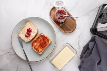 Top view of two toasts with butter and strawberry jam on gray plate, with knife, butter, jam jar,...