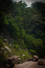 Tropical mountain road surrounded by dense jungle and rocky slopes under dramatic cloudy sky.
