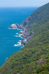 Scenic tropical coastline with steep rocky cliffs and lush green forest descending into turquoise ocean waters. 