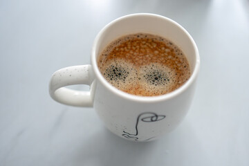 A top-down view of a cup of coffee with a creamy froth pattern that resembles a pair of wide, cartoon-like eyes. The white ceramic mug sits on a light, smooth surface.