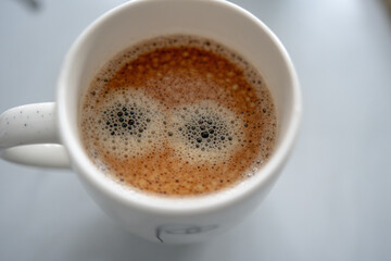 A top-down view of a cup of coffee with a creamy froth pattern that resembles a pair of wide, cartoon-like eyes. The white ceramic mug sits on a light, smooth surface.