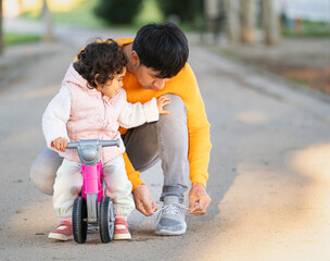Hispanic father tying his shoelaces while his daughter sits on a balance bike, enjoying a playful moment in the park