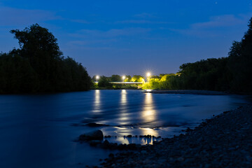 A peaceful night scene of a calm river reflecting blue twilight and street lights from a distant bridge, surrounded by trees. Long exposure captures the serene ambiance of the evening