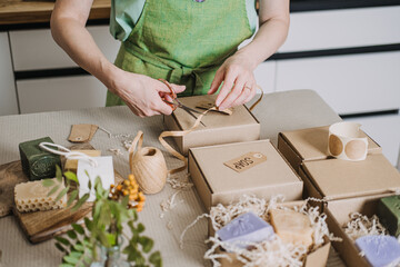 Person arranges kraft soap boxes on a table with shredded filler and handmade product tags. Handmade movement, analog creativity, DIY packaging, human touch in business