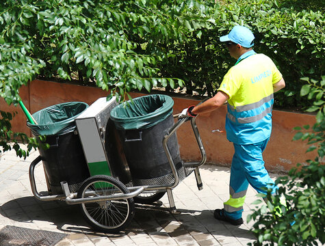 Barrendero municipal limpiando con carro, cubo y bolsas junto a jard&iacute;n y naturaleza en las calles de Madrid