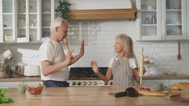 Funny old man and woman dancing in kitchen and singing at rolling-pin and spoon. Cheerful married couple having fun at home in weekend, good mood and feeling, be positive and healthy in old age