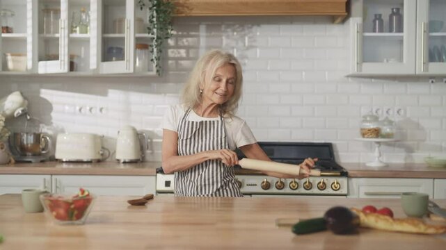 Portrait of mature housewife dancing with rolling-pin in kitchen, good mood. Cheerful grandmother enjoying favorite music and singing alone at home, do housework, cooking and feeling good, have fun