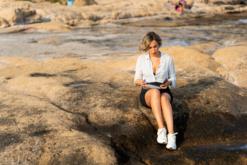 Businesswoman working remotely on rocky beach using mobile phone and notepad