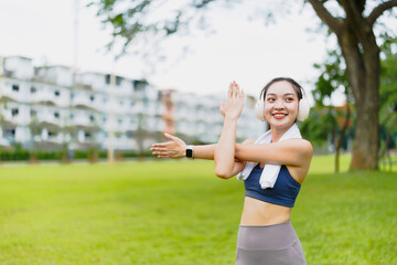 Young sportswoman stretching her arms in a park, smiling and enjoying her workout routine while listening to music through headphones, embodying a healthy and active lifestyle