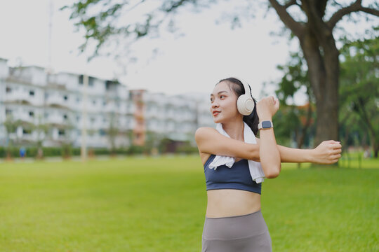 Young asian sportswoman wearing wireless headphones and smartwatch stretching arms before training in a park in the city, enjoying music and healthy lifestyle