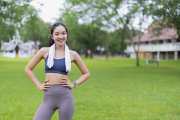Fototapeta premium Young Asian sportswoman wearing stylish sportswear and a smartwatch, smiling confidently while posing with hands on hips in a vibrant green park, embodying a healthy, active lifestyle