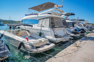 Several luxury yachts and boats are moored in a sunny marina with clear water, surrounded by calm...