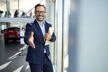 Confident male car salesman in a formal suit standing inside a modern and bright dealership showroom, smiling and reaching out to shake hands with a potential customer, offering a warm welcome.